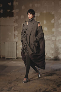 Model walking in a gray, sculptural coat with shoulder cutouts, floral appliques, and a burgundy draped cape in an industrial studio setting.