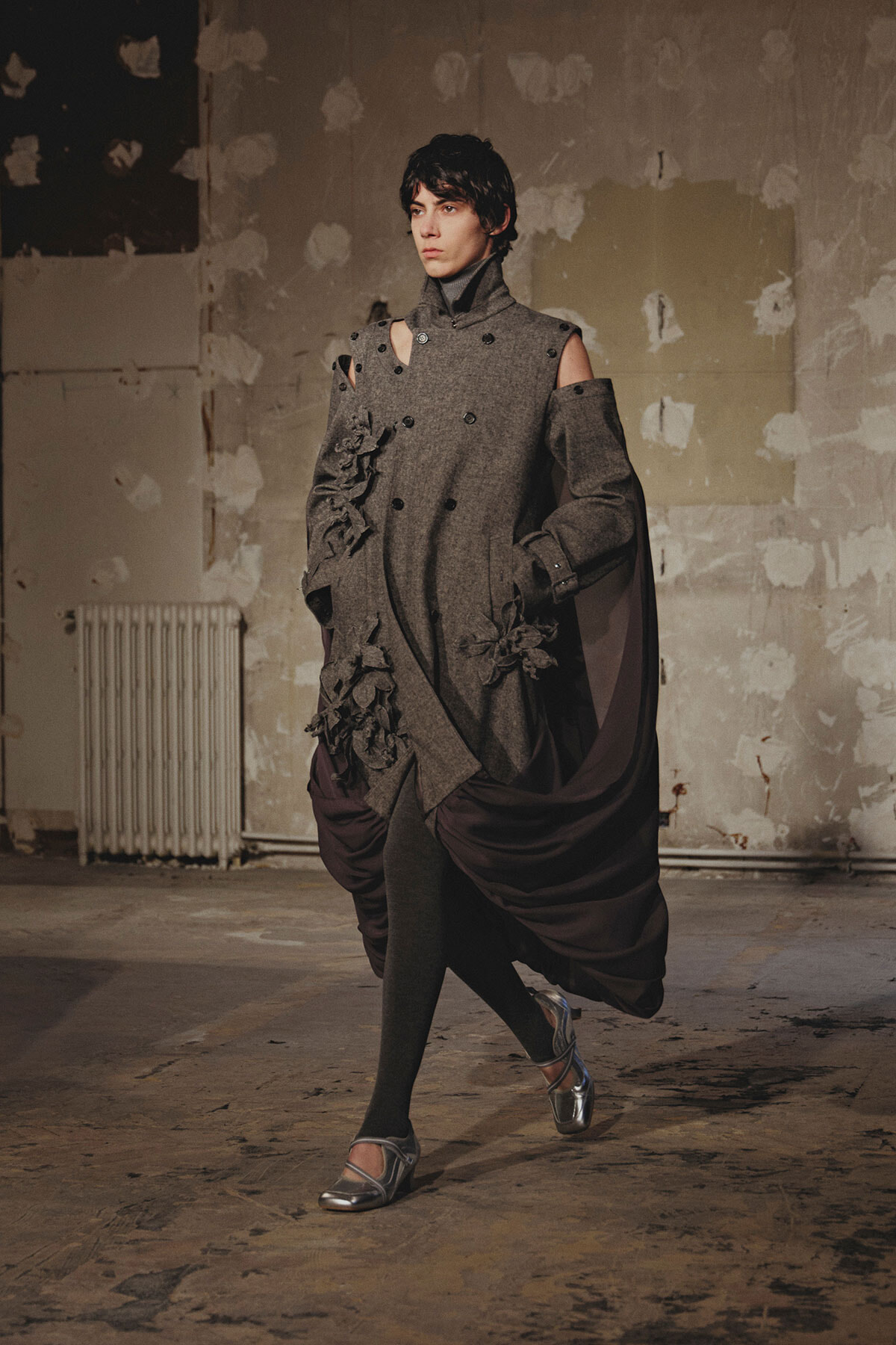 Model walking in a gray, sculptural coat with shoulder cutouts, floral appliques, and a burgundy draped cape in an industrial studio setting.