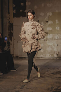 Model walks a runway in a beige sculptural outfit with layered petal-like ruffles, black tights, and pale flats in an industrial backdrop.