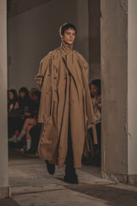 Model walks the runway in a long oversized beige trench coat with layered detailing, an industrial venue backdrop and seated audience in the background.