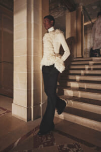 Person wearing a cream fringe top over a white turtleneck and black pants, posing on a marble staircase in a grand interior.