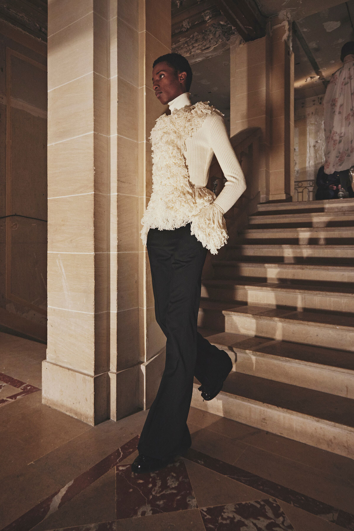 Person wearing a cream fringe top over a white turtleneck and black pants, posing on a marble staircase in a grand interior.