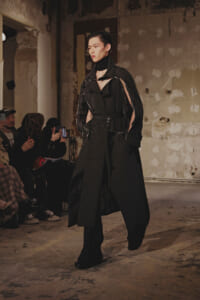 Model walking a black, oversized trench on a runwayshow in an industrial space; audience watches and photographs on the left.