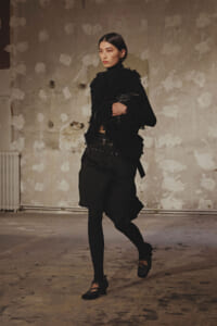 Model walking in a black textured outfit with fringe details, belt, and Mary Jane shoes in an industrial studio setting.