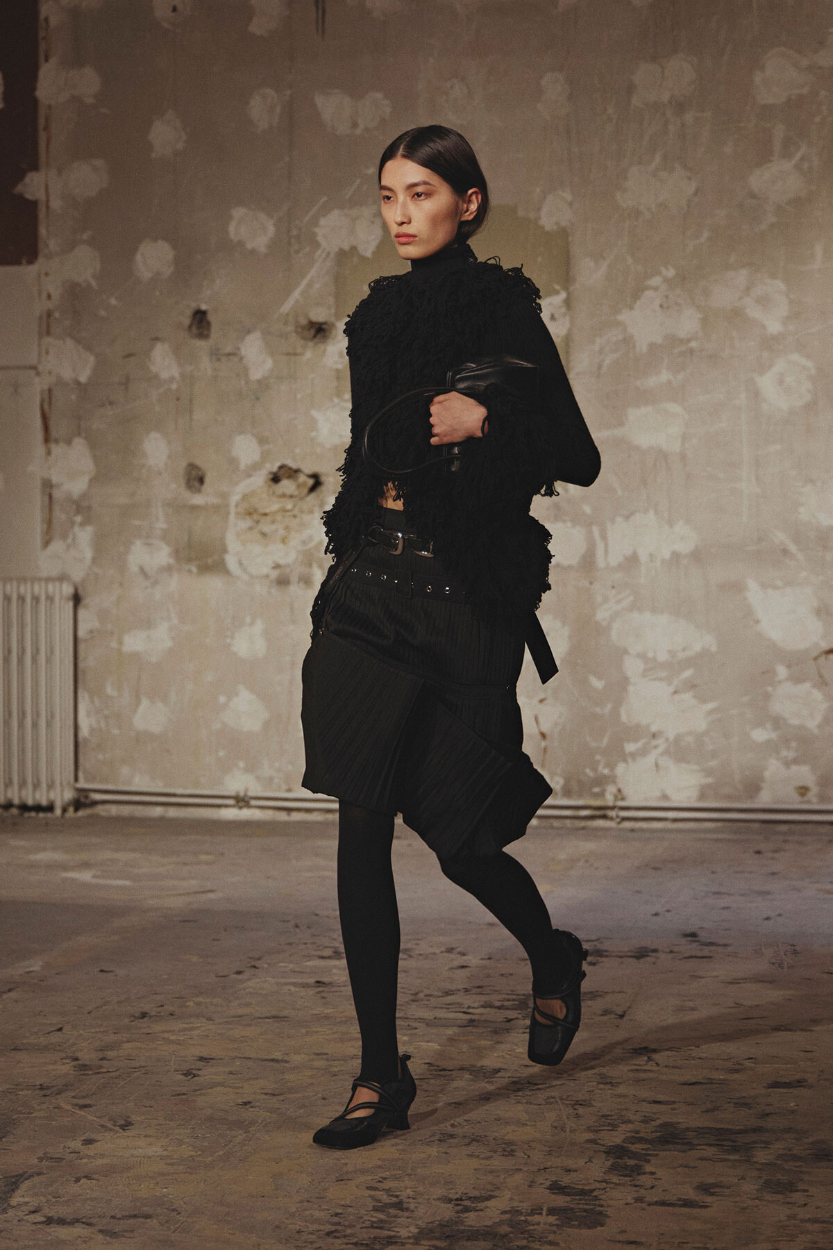 Model walking in a black textured outfit with fringe details, belt, and Mary Jane shoes in an industrial studio setting.