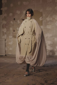 Model wearing a beige double-breasted trench with a flowing cape-like overlay and gathered hem, standing in a studio.