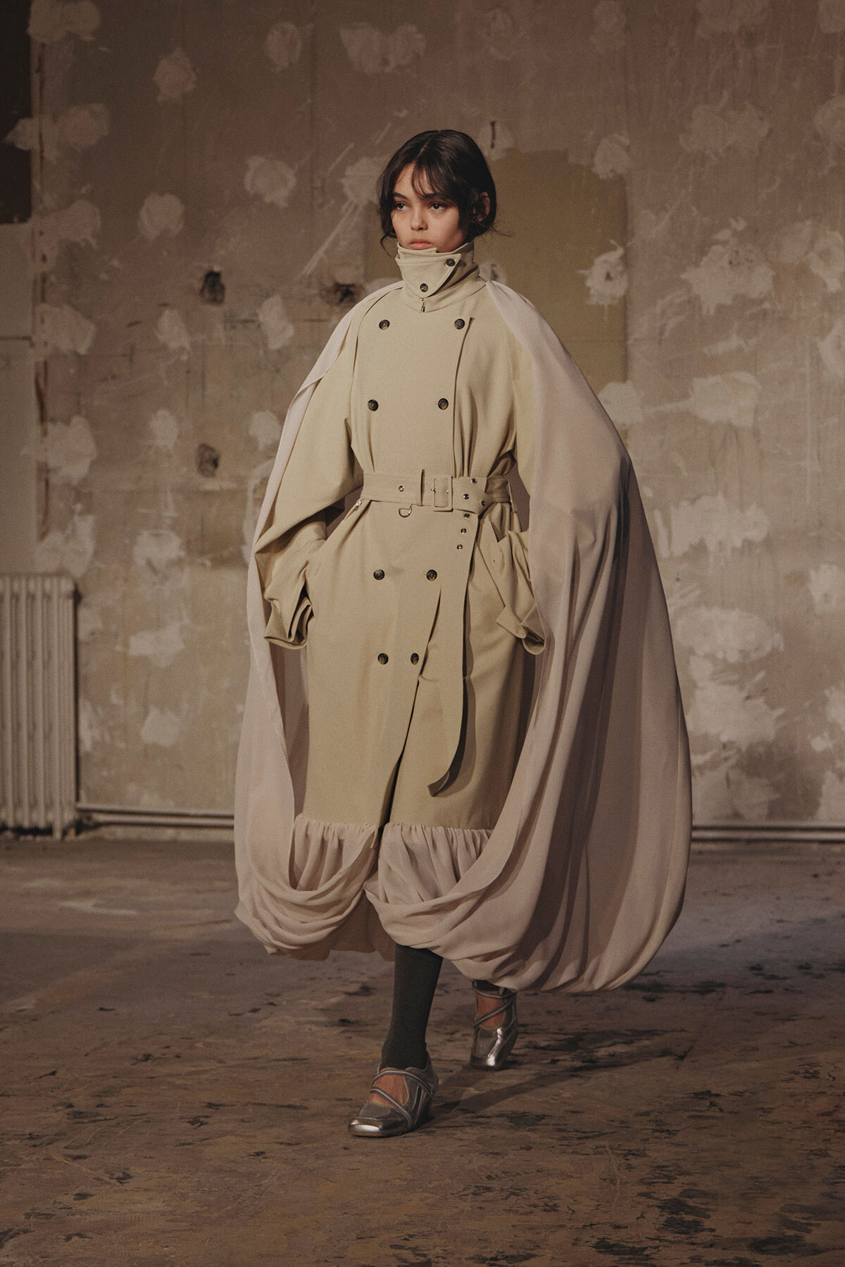 Model wearing a beige double-breasted trench with a flowing cape-like overlay and gathered hem, standing in a studio.