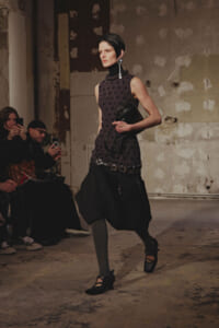 Model walking a runway in a sleeveless black polka-dot top and a voluminous black pleated skirt, clutch in hand.