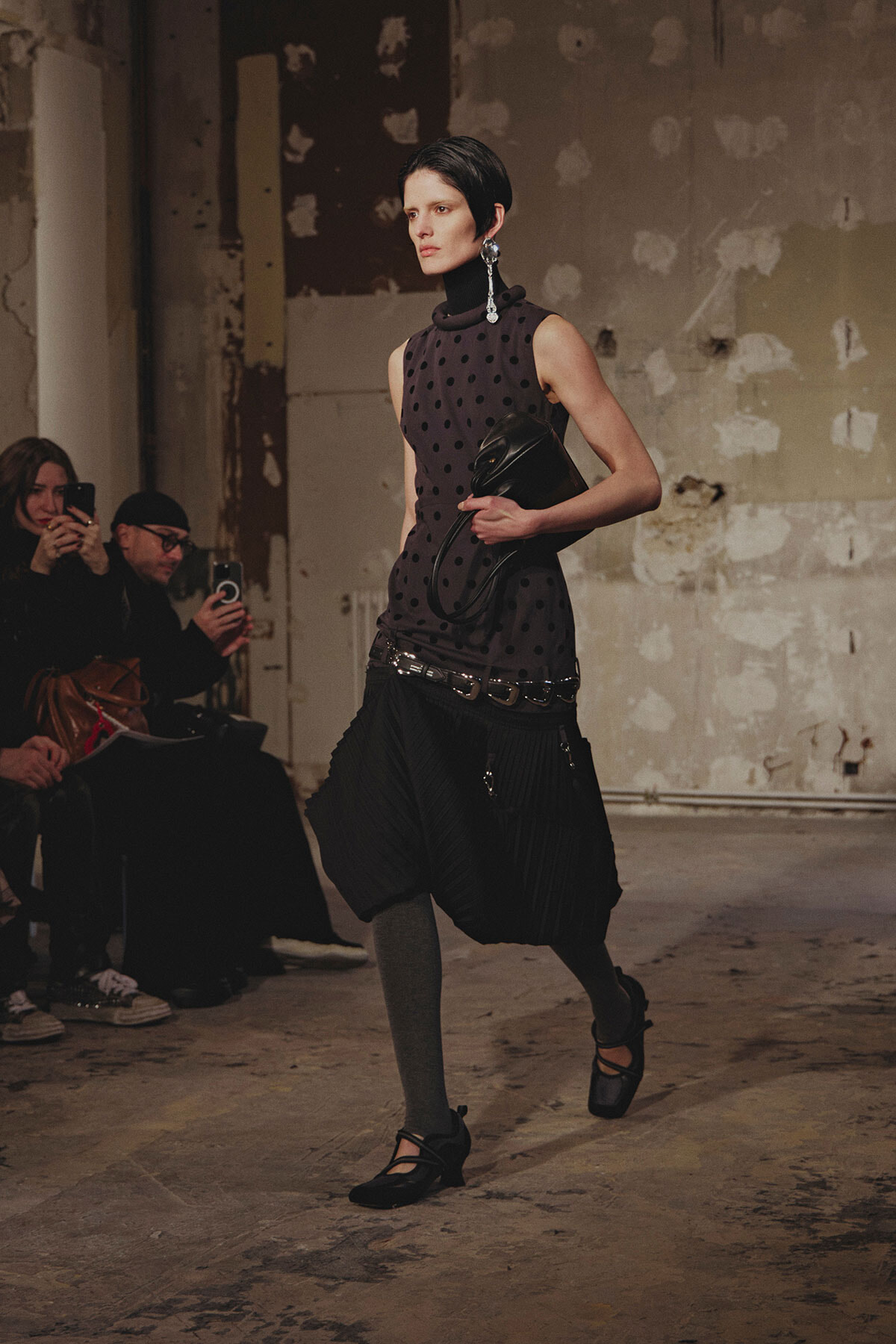 Model walking a runway in a sleeveless black polka-dot top and a voluminous black pleated skirt, clutch in hand.