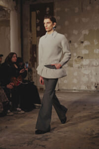Model walks a runway in a light gray oversized knit top with a layered peplum detail and wide gray trousers, industrial backdrop behind.