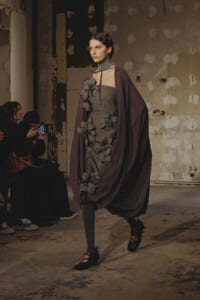 Model walks a fashion runway in a textured gray strapless dress with dark floral appliqués and a deep purple cape in an industrial setting