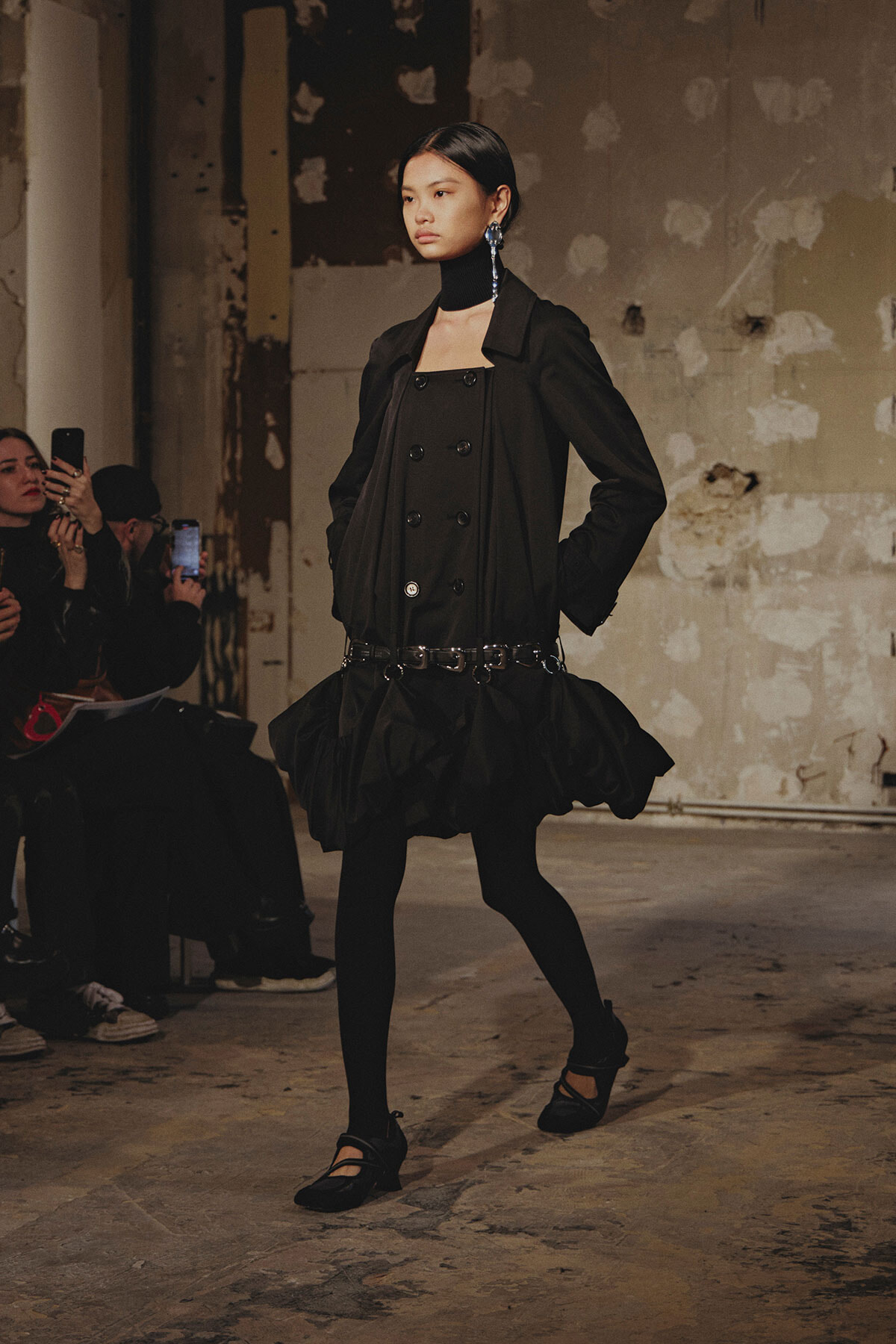 Model walks a fashion show in an all-black layered ensemble with a double-breasted top, flared skirt, and chunky belt, wearing black tights and Mary Jane shoes in an industrial setting.