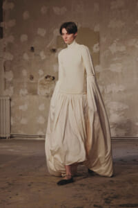 Model wearing an ivory high-neck, long-sleeved gown with a voluminous gathered skirt in a bare, textured studio.