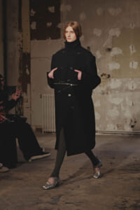 Model walking a fashion show in an industrial venue, wearing a long black double-breasted coat with a belt and metallic flats.