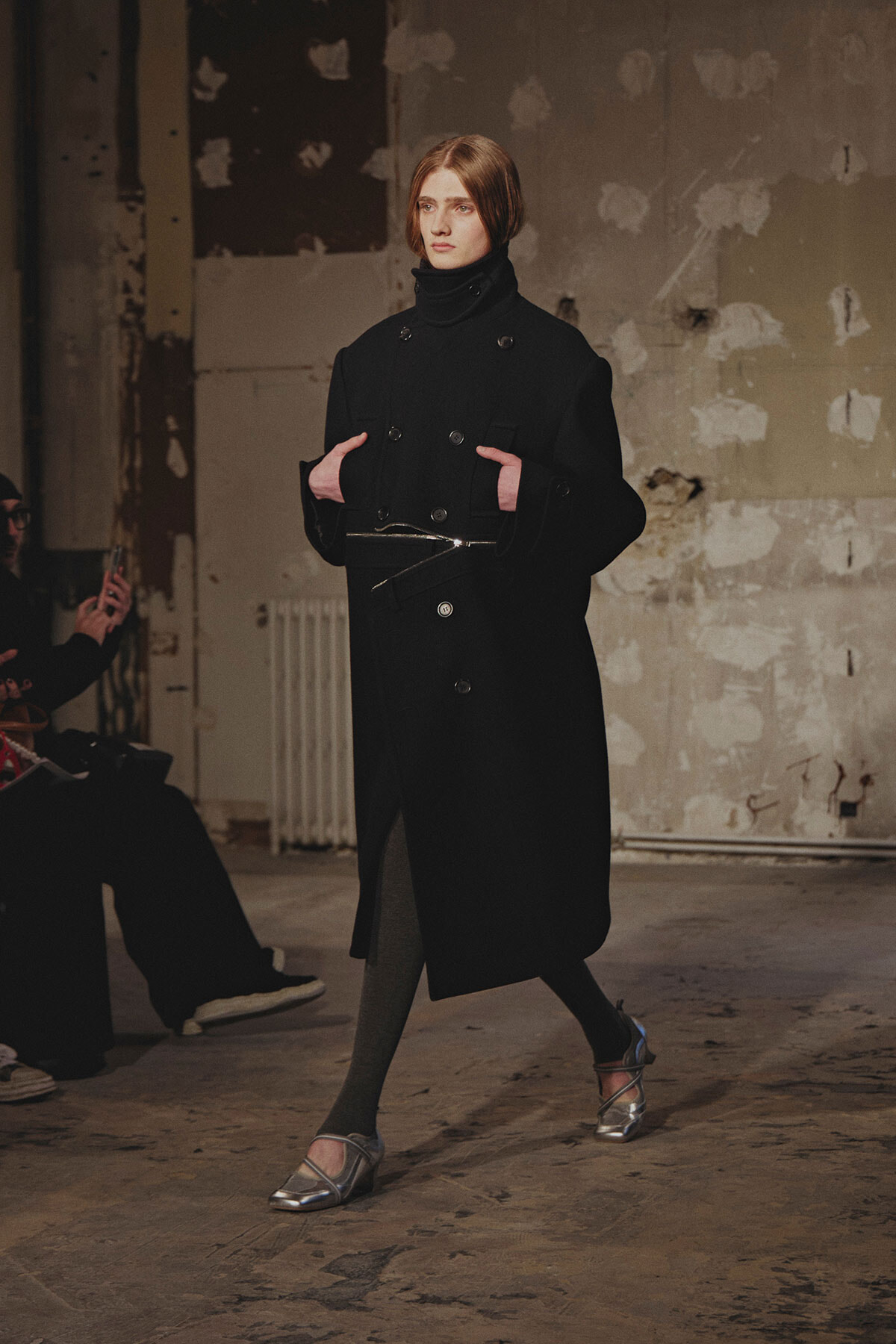 Model walking a fashion show in an industrial venue, wearing a long black double-breasted coat with a belt and metallic flats.