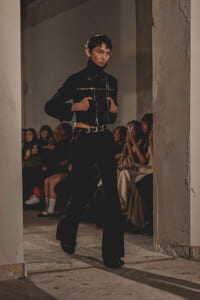 Model walking a runway in a dark, cropped black jacket with metallic details; audience sits in the background, watching the show in an industrial venue.