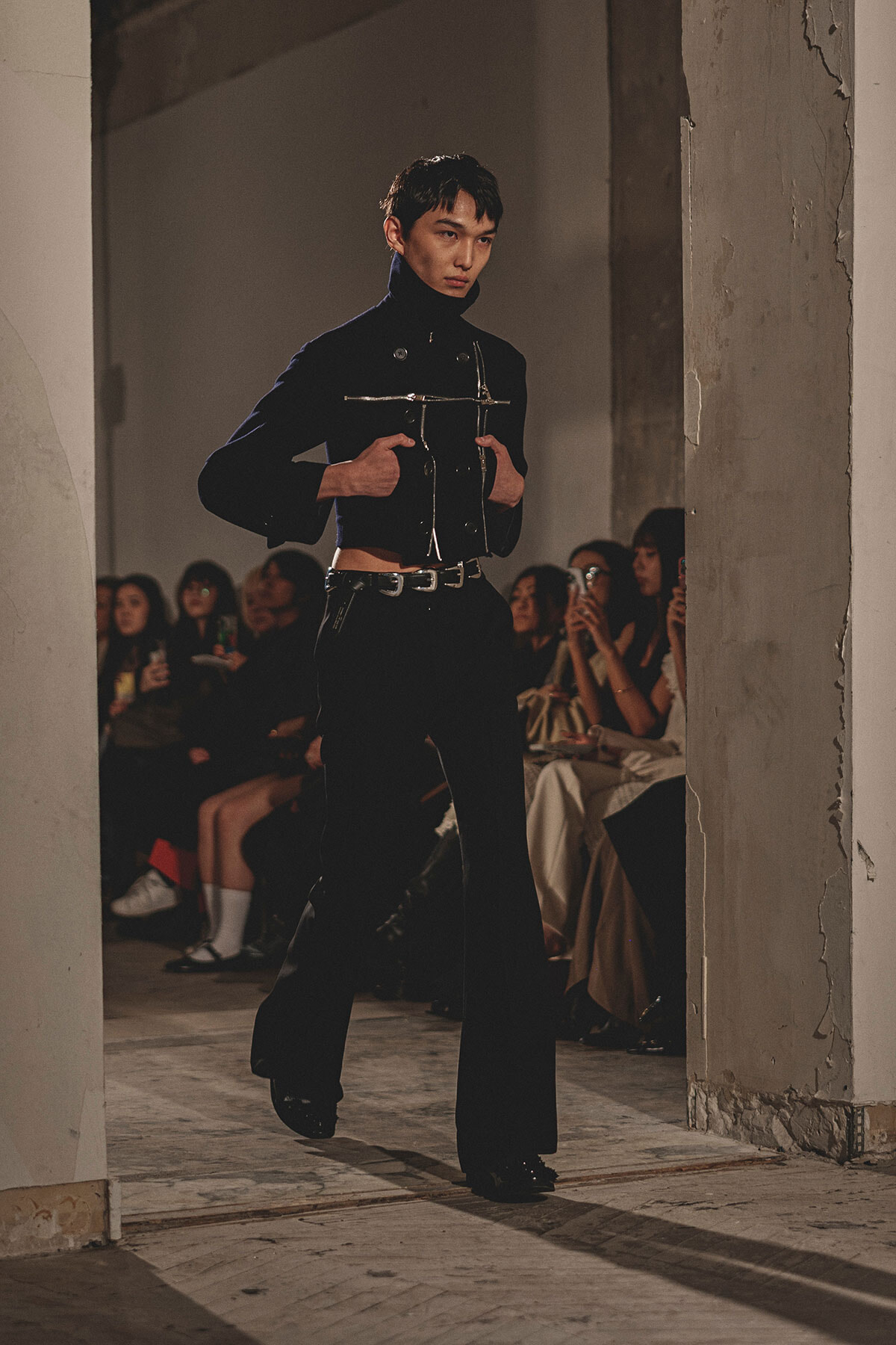 Model walking a runway in a dark, cropped black jacket with metallic details; audience sits in the background, watching the show in an industrial venue.