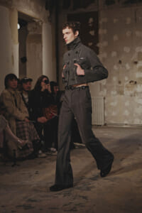 Male model in a gray high-collar jacket and wide-legged trousers walks a runway in a raw, industrial space with spectators seated along the side.