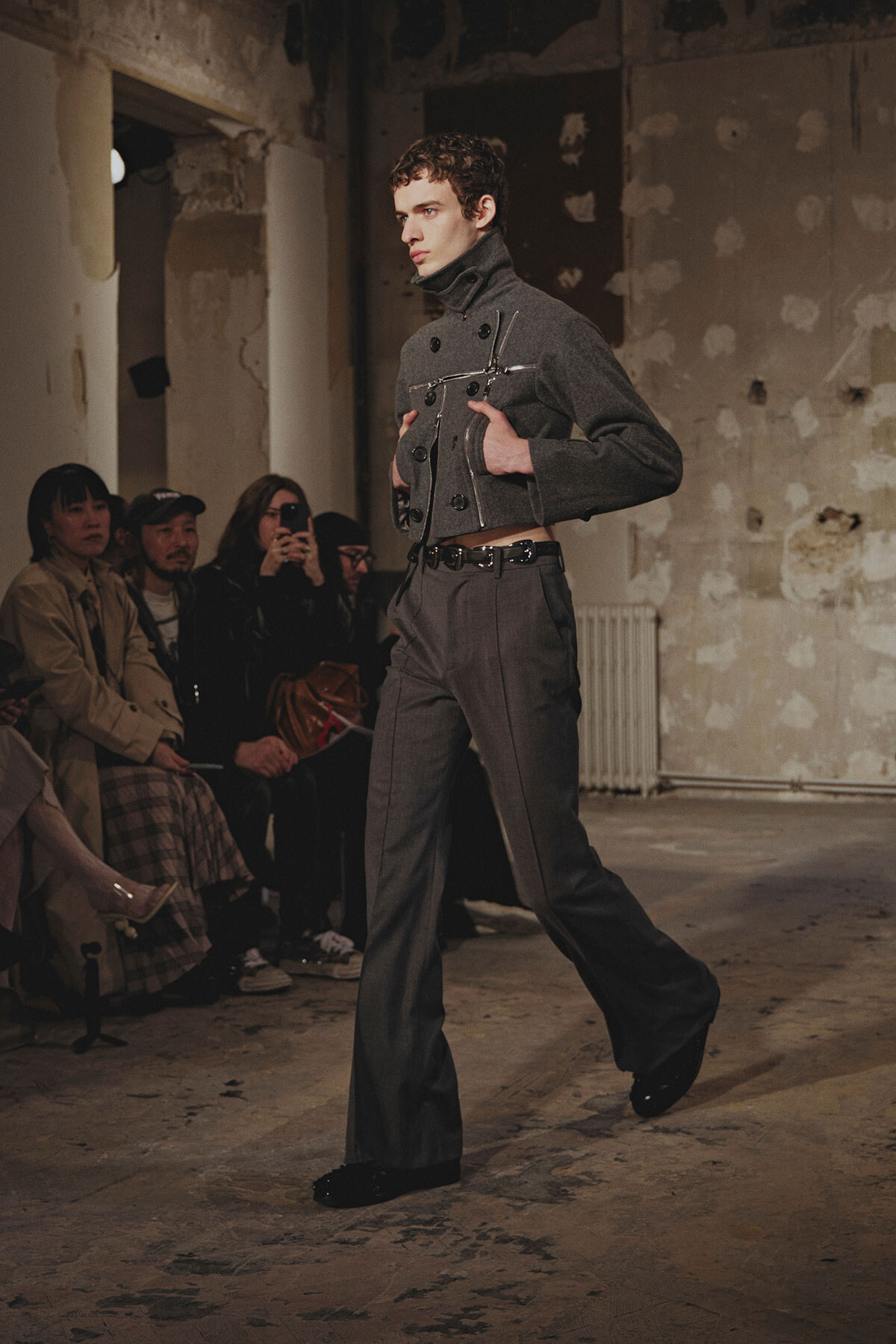 Male model in a gray high-collar jacket and wide-legged trousers walks a runway in a raw, industrial space with spectators seated along the side.