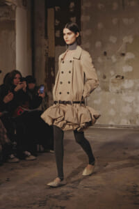 Model walks the runway in a beige trench-inspired ensemble with a double-breasted bodice and a voluminous bubble skirt, black tights, and beige flats. Audience members in the background.