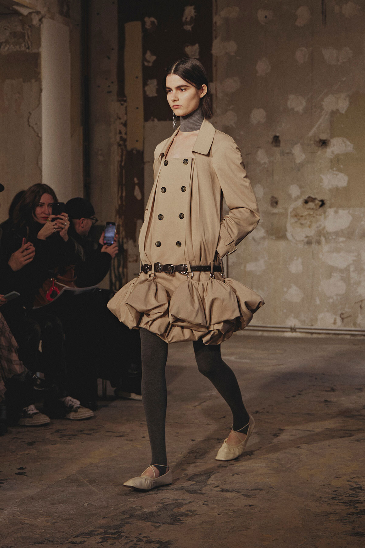Model walks the runway in a beige trench-inspired ensemble with a double-breasted bodice and a voluminous bubble skirt, black tights, and beige flats. Audience members in the background.