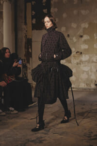 Model walks the runway in an all-black polka-dot ensemble with a high collar and voluminous skirt in an industrial, unfinished venue.