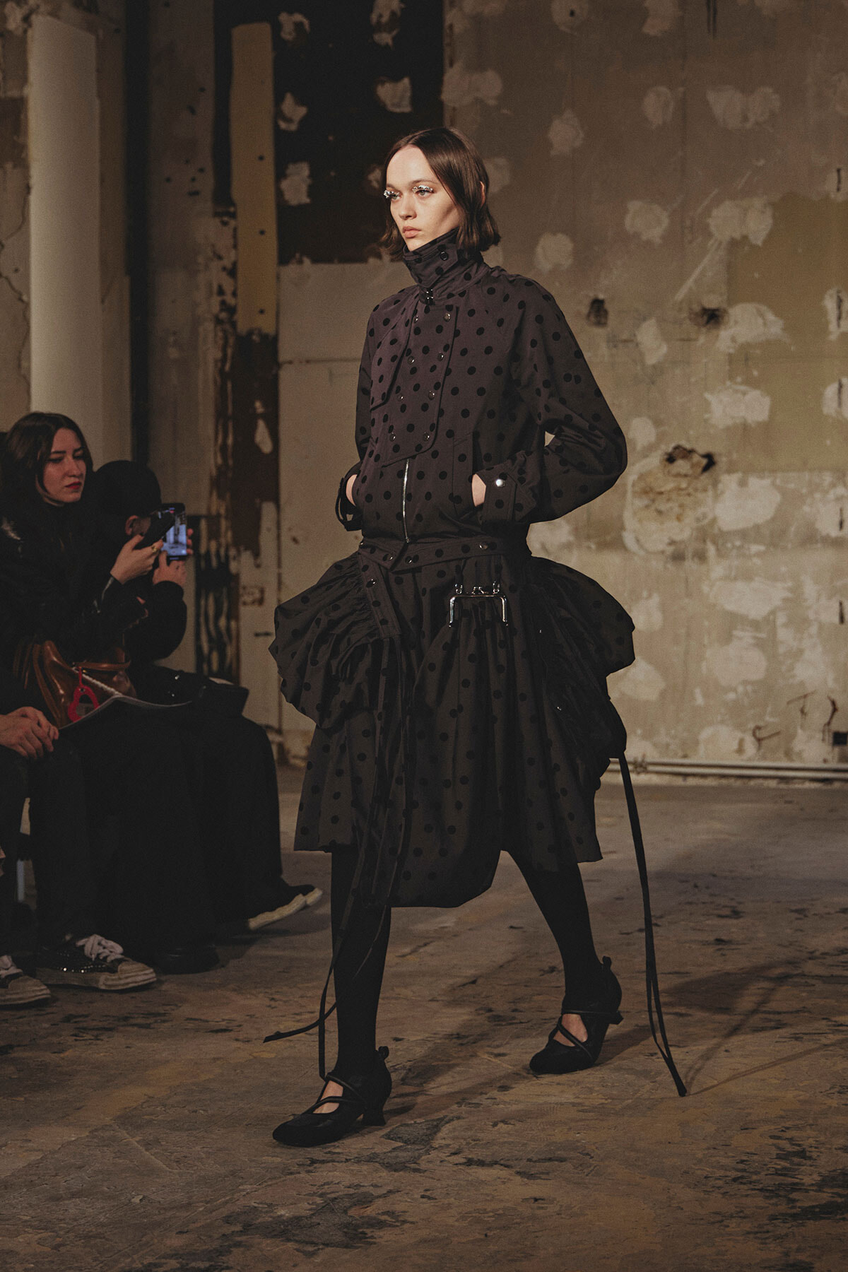 Model walks the runway in an all-black polka-dot ensemble with a high collar and voluminous skirt in an industrial, unfinished venue.