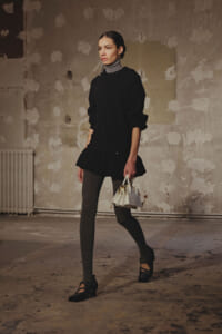 Model walking in a dim studio wearing a layered black knit dress over a gray turtleneck, gray tights, and black Mary Jane shoes, carrying a small white handbag with gold hardware.