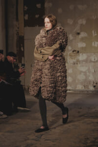 Model walks a fashion show in a large taupe ruffled coat, paired with gray tights and black flats, on an industrial backstage runway.