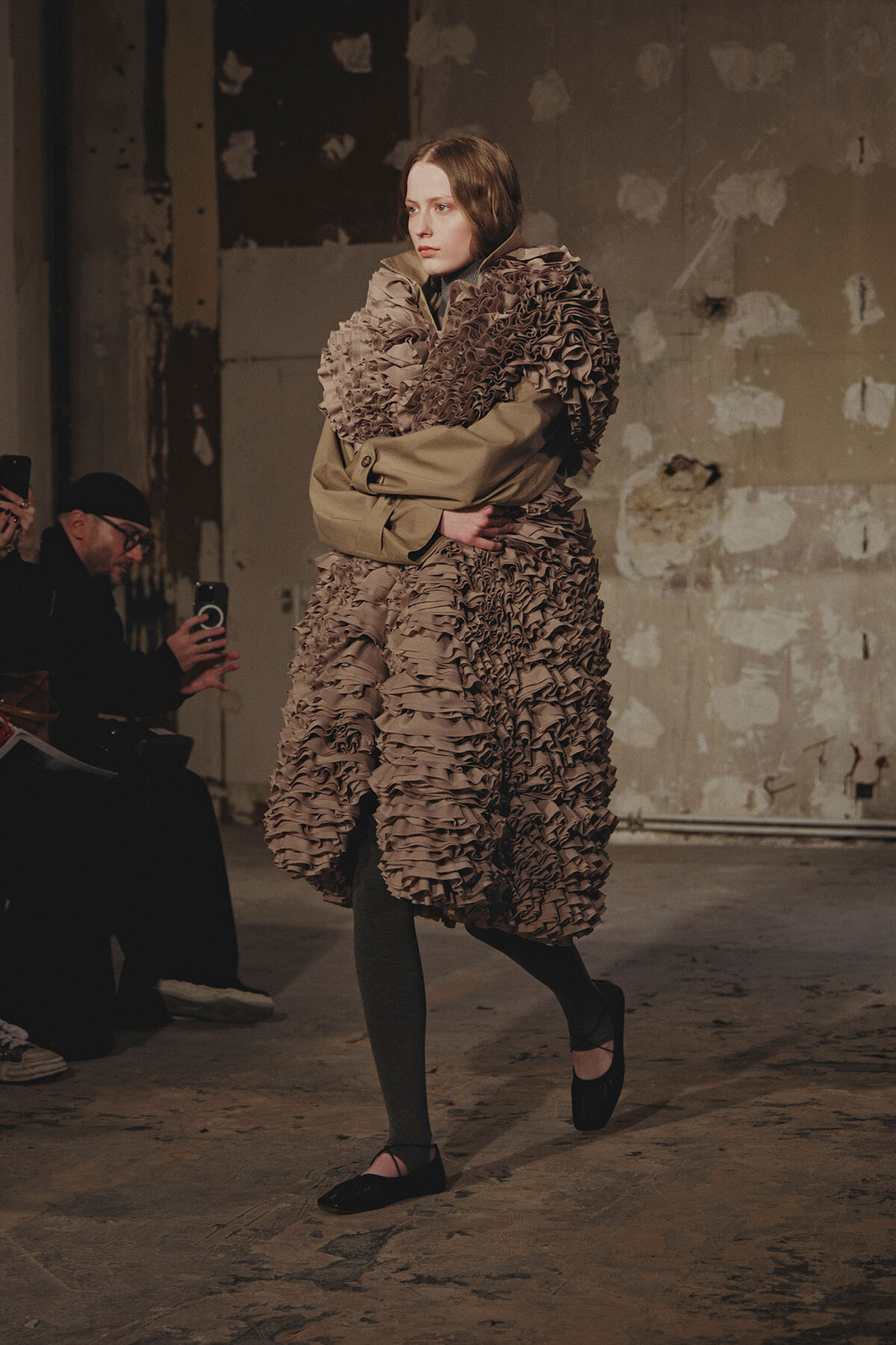 Model walks a fashion show in a large taupe ruffled coat, paired with gray tights and black flats, on an industrial backstage runway.