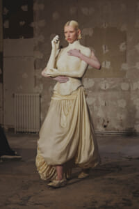 Person in a cream gown cradles a white sculpture arm across the chest in a gritty, unfinished studio.