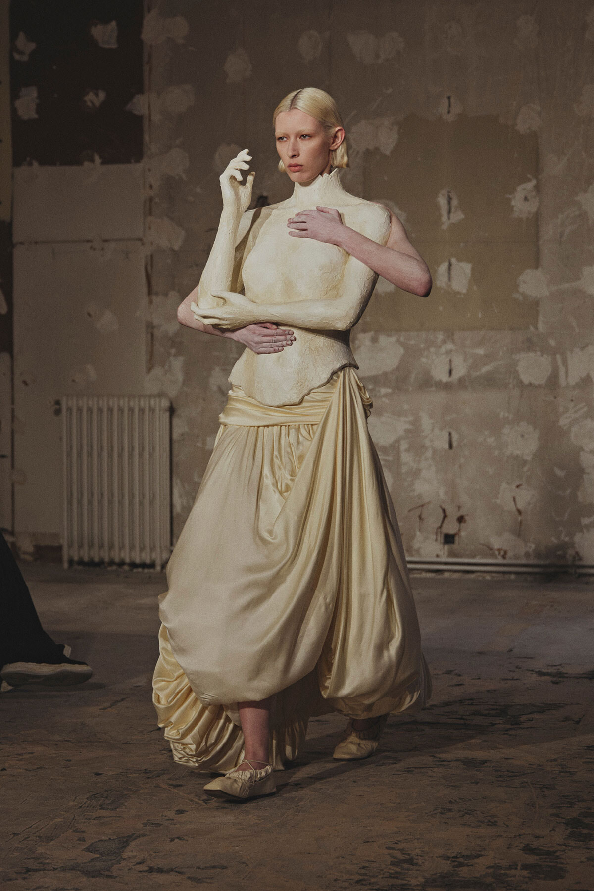 Person in a cream gown cradles a white sculpture arm across the chest in a gritty, unfinished studio.