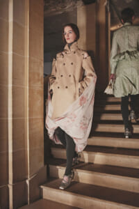 Model in a beige double-breasted trench coat descends a stone staircase, with a pink floral scarf flowing from the coat.