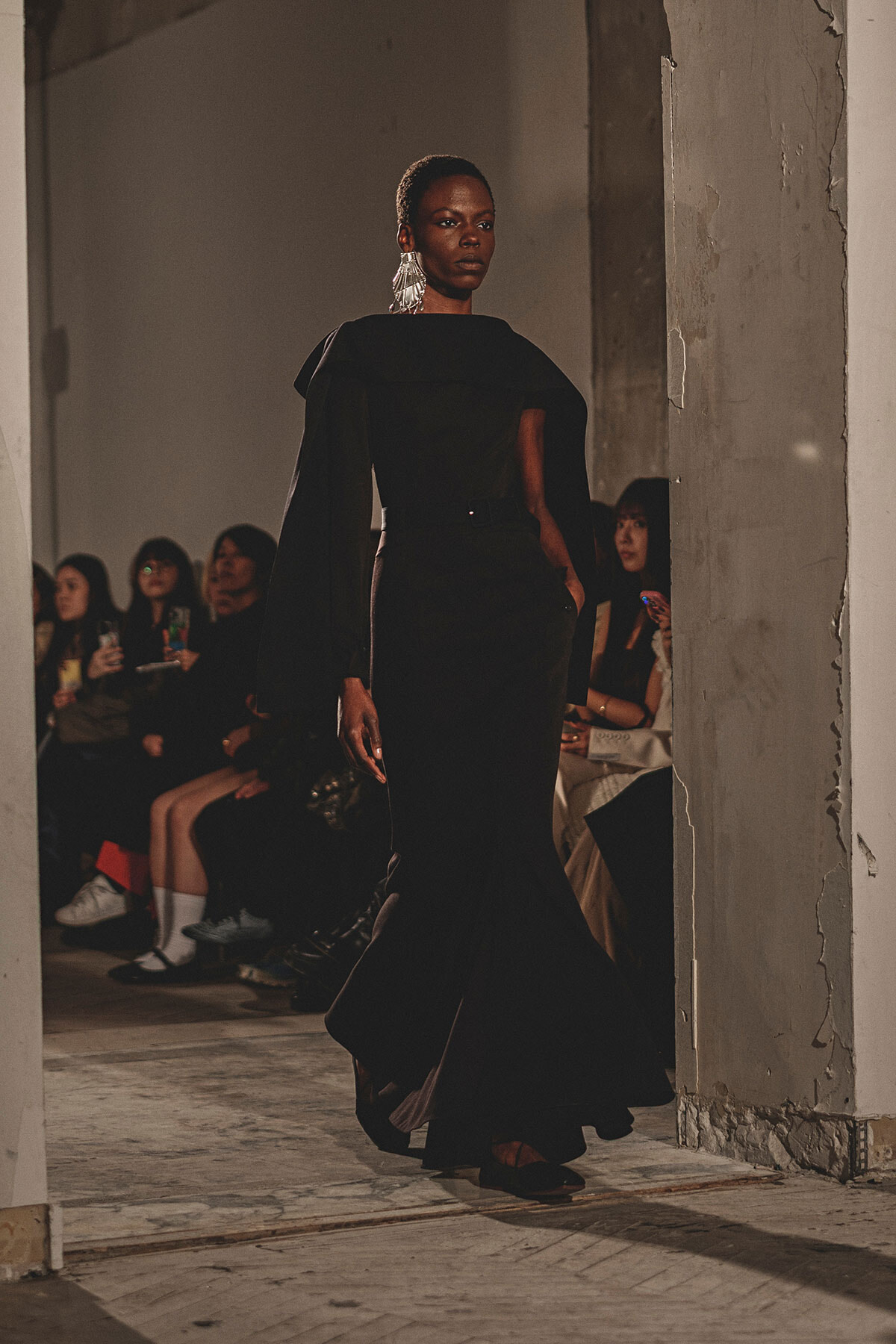 Model walking the runway in a black, cape‑shouldered gown with large silver earrings, audience in the background.