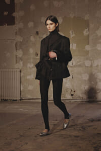 Woman in a black tailored suit with a high-neck top, standing in a dilapidated studio with peeling walls and a radiator in the background.