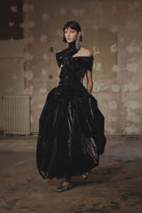 Model in a dramatic black satin off-shoulder gown with a voluminous skirt, walking in a gritty, unfinished studio.