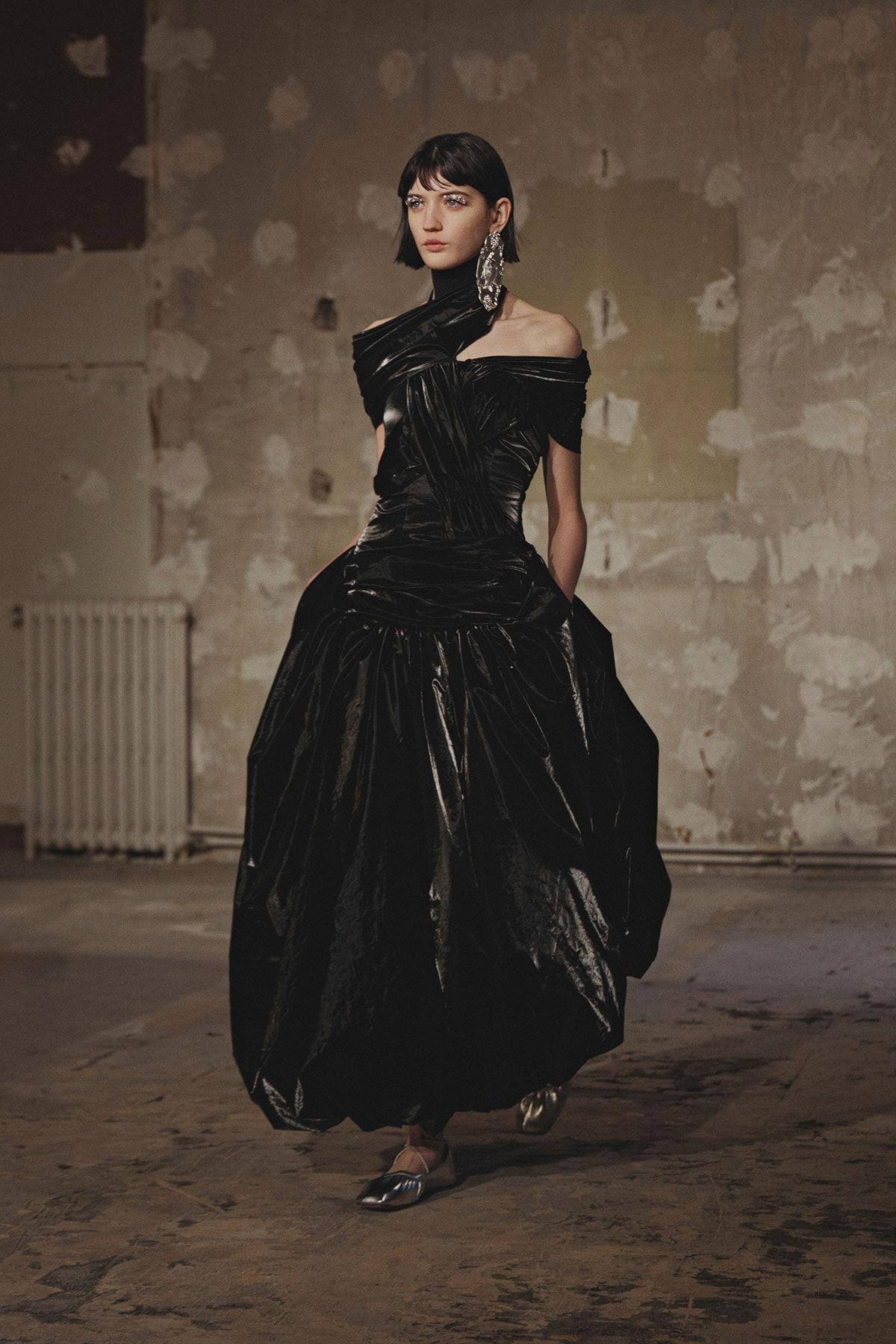 Model in a dramatic black satin off-shoulder gown with a voluminous skirt, walking in a gritty, unfinished studio.