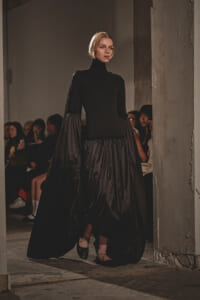 Model walks the runway in a black turtleneck with a dramatic, voluminous skirt as spectators watch in a dim, industrial setting.