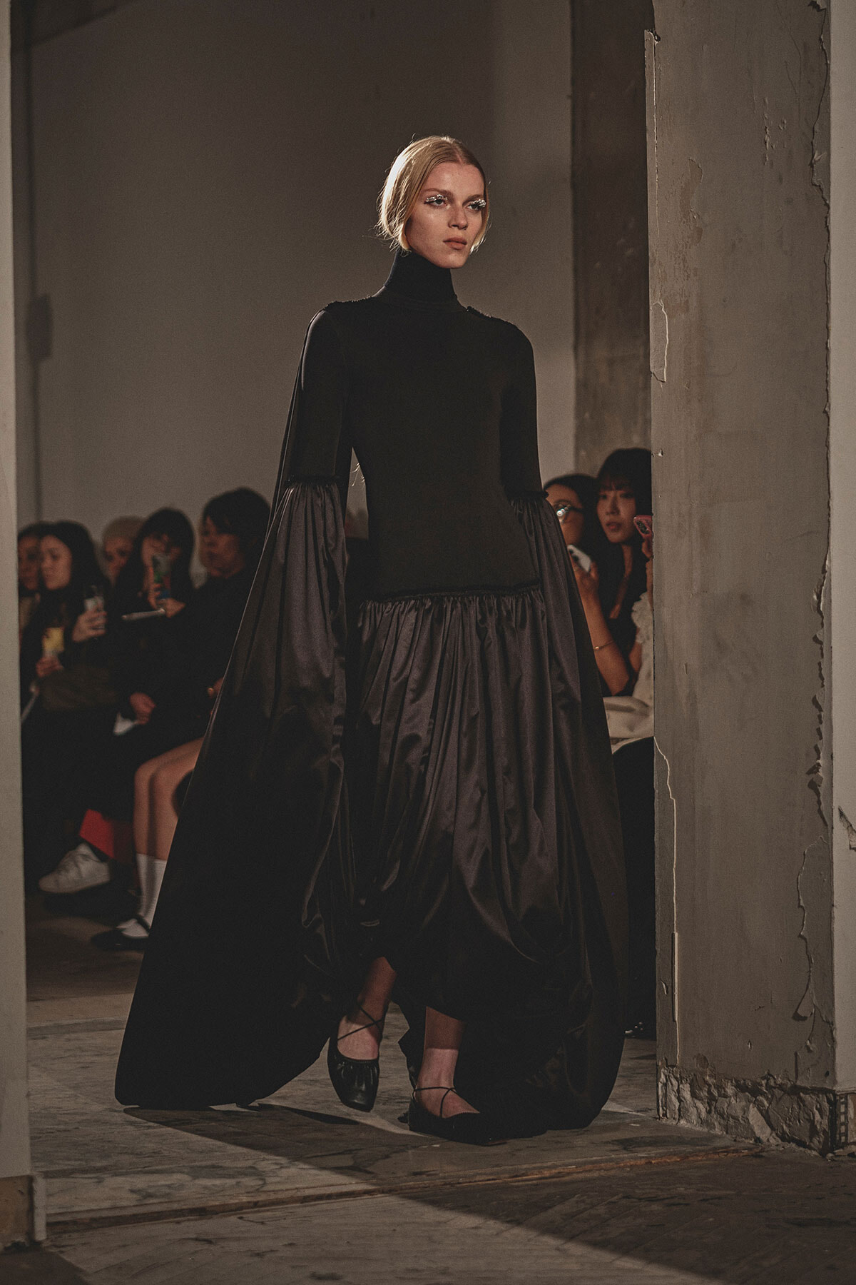 Model walks the runway in a black turtleneck with a dramatic, voluminous skirt as spectators watch in a dim, industrial setting.