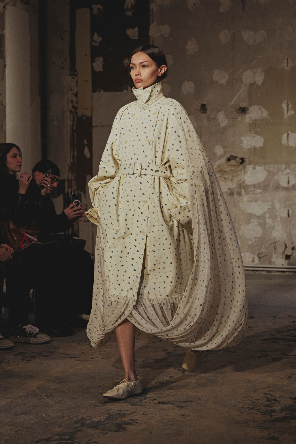 Model on a runway wearing a voluminous cream cape-dress with a dotted pattern and belt, performing in an industrial setting.