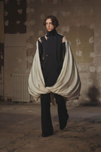 Model wearing a black double-breasted outfit with shoulder cutouts, wrapped in a flowing ivory cape in a bare industrial studio.