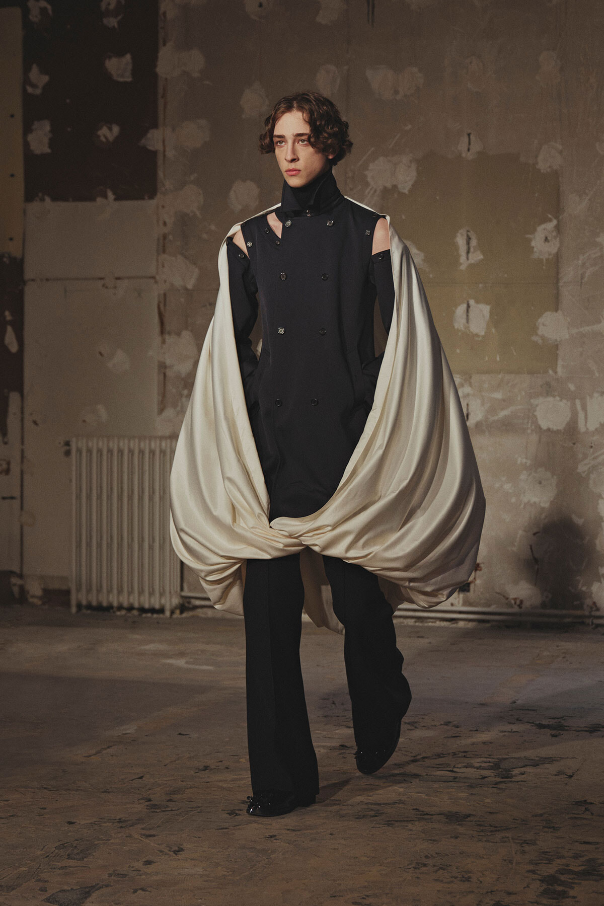 Model wearing a black double-breasted outfit with shoulder cutouts, wrapped in a flowing ivory cape in a bare industrial studio.