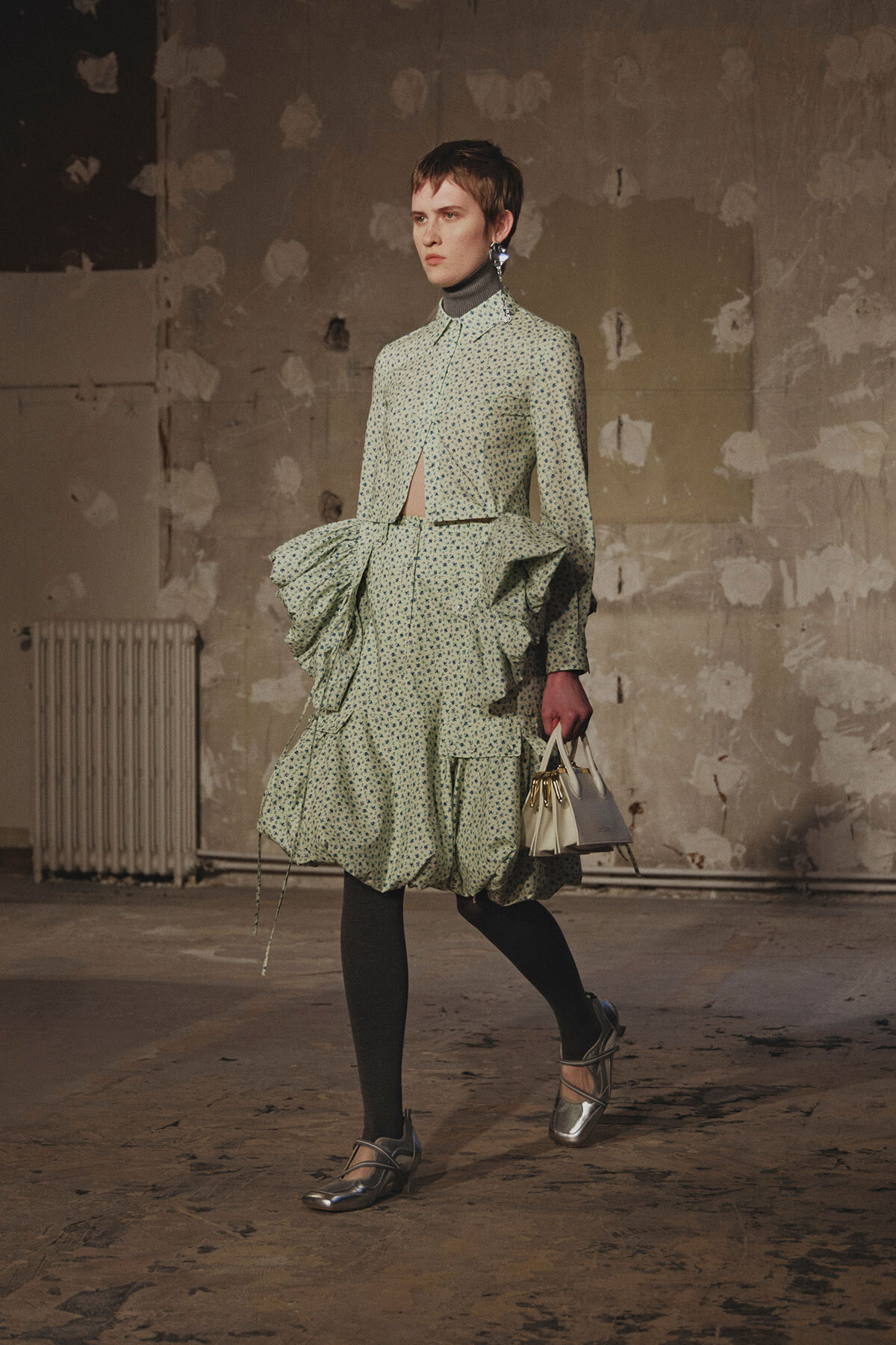 Model walking in an avant-garde green printed dress with large structured ruffles, gray tights, and metallic shoes, carrying a white handbag in an industrial setting