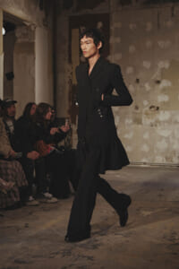 Model in a black, double-breasted coat with button detailing walks a runway in an industrial, unfinished venue with seated spectators nearby.