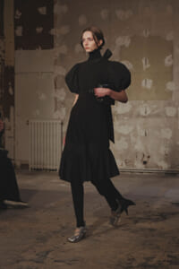 Woman in a black puff-sleeve dress walks through a distressed, industrial-style space, holding a small black clutch.