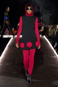 Model walks a fashion runway in a red-and-black futuristic outfit: red leggings and hoodie under a black sleeveless dress with large red circular patches at the hem, plus a visor.