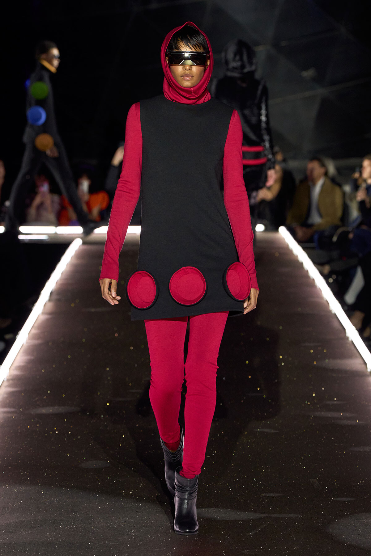 Model walks a fashion runway in a red-and-black futuristic outfit: red leggings and hoodie under a black sleeveless dress with large red circular patches at the hem, plus a visor.