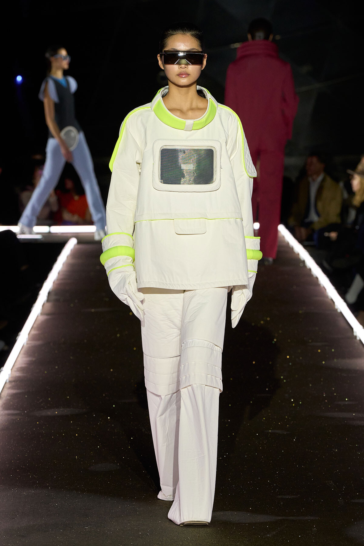 Model on a runway wearing a futuristic white spacesuit-inspired outfit with neon yellow trim and oversized visor sunglasses, mid-stride.
