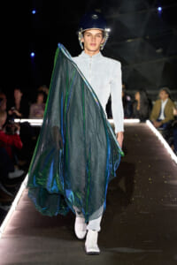 Model on a fashion runway wearing a metallic teal cape over a light gray zippered outfit and a dark helmet, white boots visible.