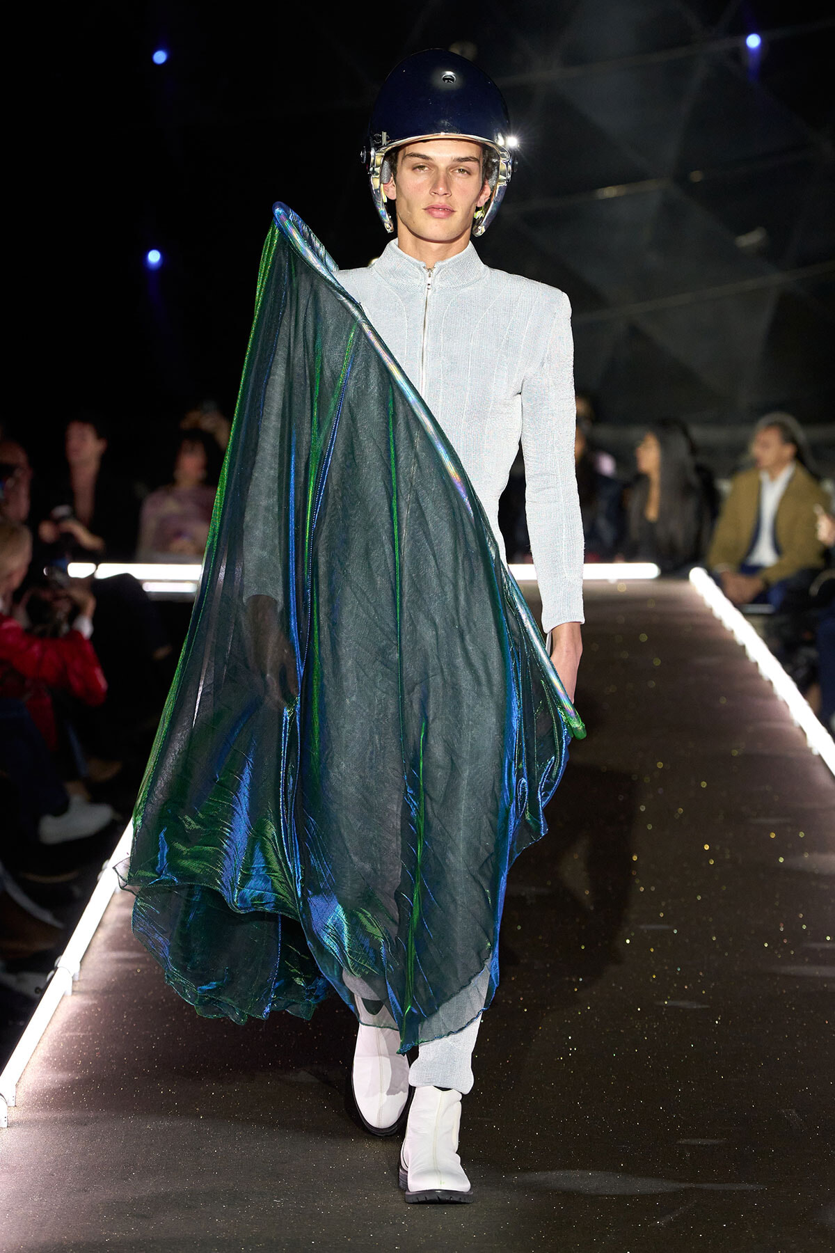 Model on a fashion runway wearing a metallic teal cape over a light gray zippered outfit and a dark helmet, white boots visible.